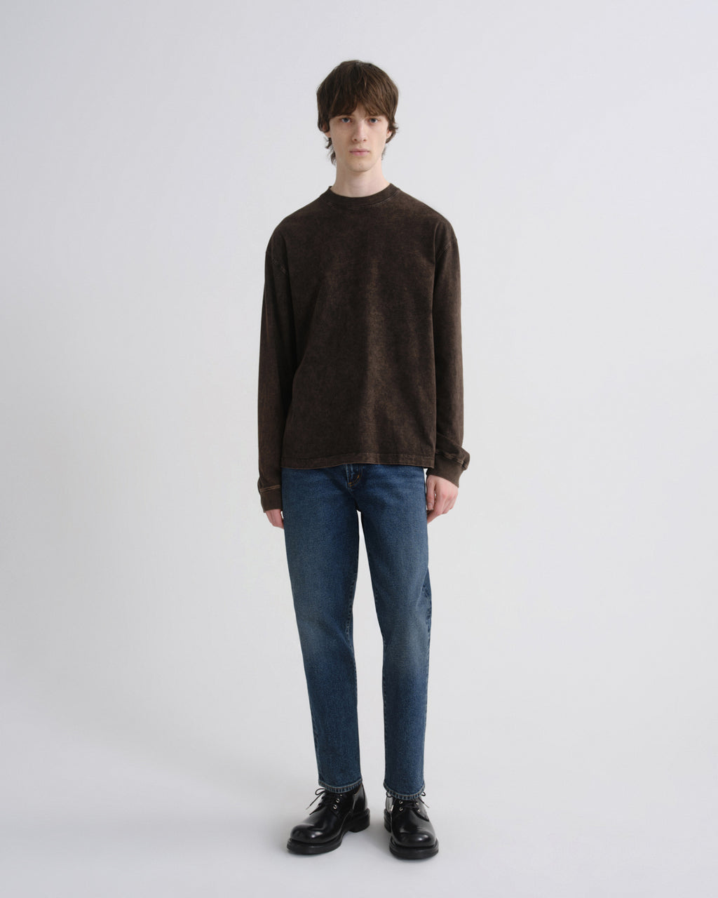 A model stands against a light gray studio background wearing a dark brown long-sleeve crewneck sweater, blue jeans, and black leather shoes.