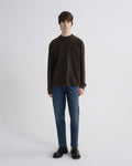 A model stands against a light gray studio background wearing a dark brown long-sleeve crewneck sweater, blue jeans, and black leather shoes.