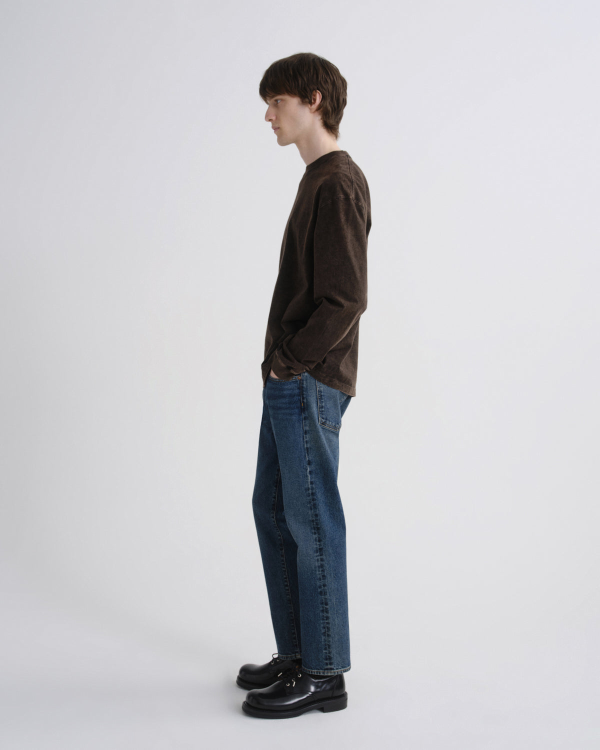 A man in profile wearing a dark brown corduroy long-sleeve shirt, blue jeans, and black leather shoes, standing against a light gray studio background.