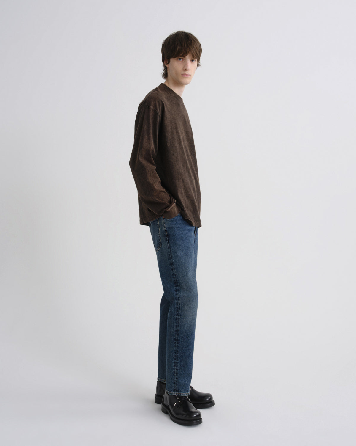 A man wearing a dark brown long-sleeve shirt, blue jeans, and black leather shoes, standing in profile against a light gray studio background.