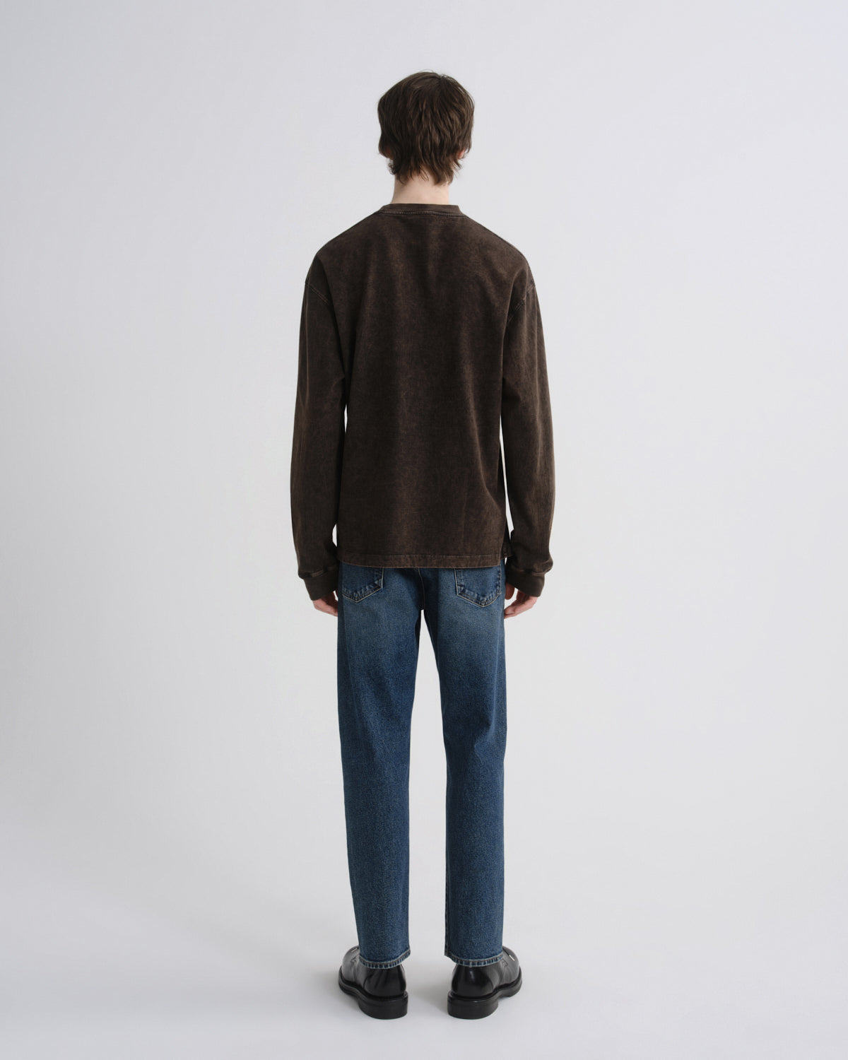 A man viewed from behind wearing a dark brown long-sleeve crewneck sweater, blue jeans, and black shoes, standing against a light gray studio background.