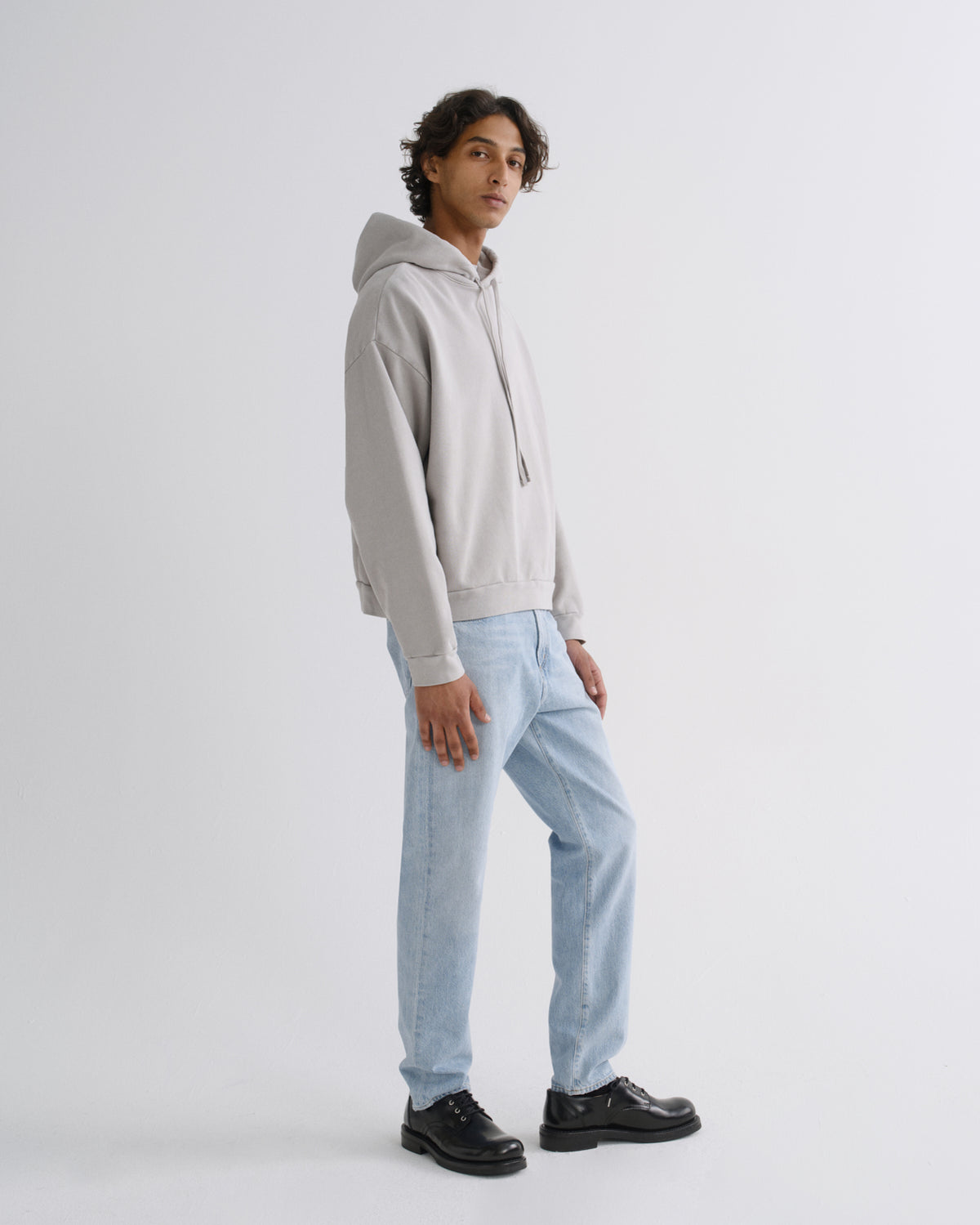 A model wearing a light gray pullover hoodie, light-wash blue jeans, and black leather shoes, standing in profile against a plain light gray studio background.