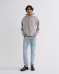 A model stands against a light gray studio background wearing a light gray oversized hoodie, light blue jeans, and black leather shoes.