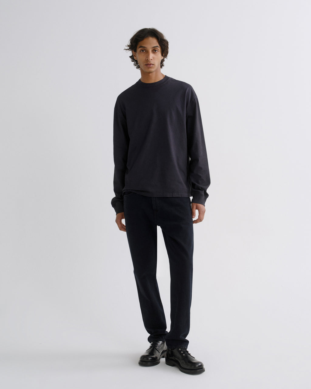 A man stands against a light gray studio background wearing a dark charcoal long-sleeve crewneck sweatshirt, dark navy pants, and black leather shoes.