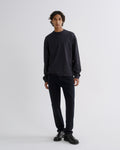 A man stands against a light gray studio background wearing a dark charcoal long-sleeve crewneck sweatshirt, dark navy pants, and black leather shoes.