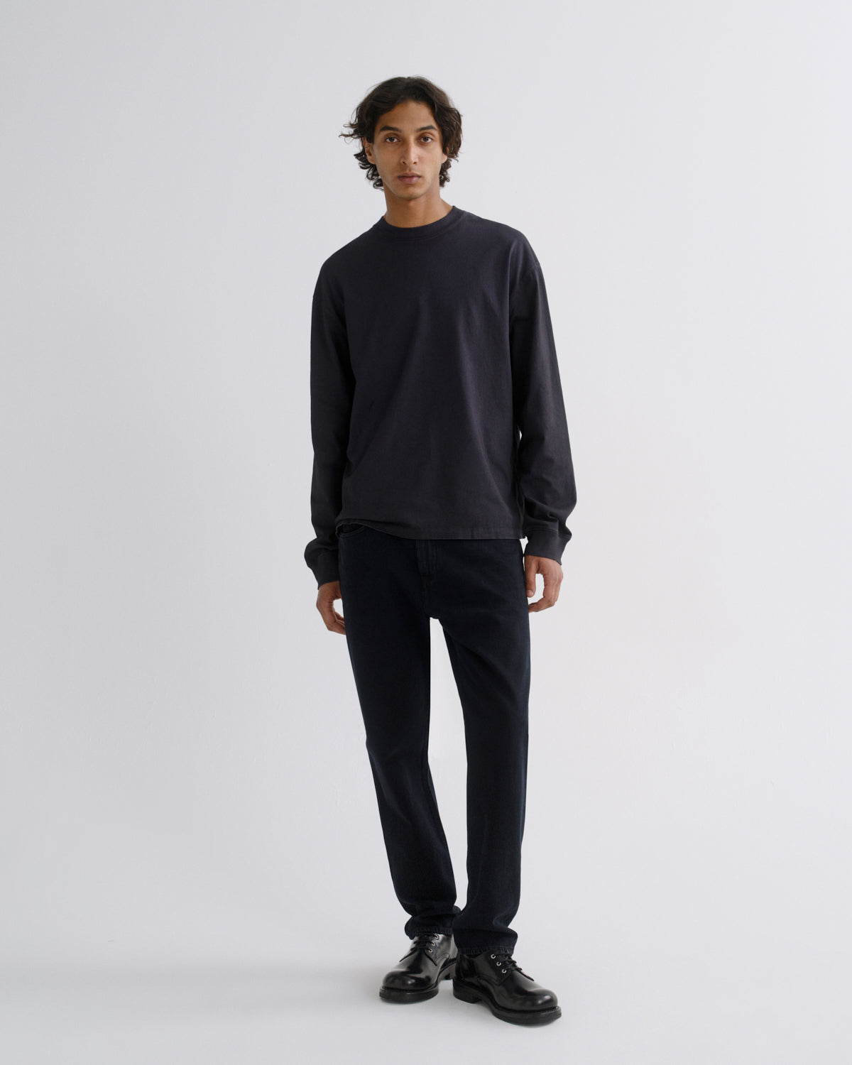 A man stands against a light gray studio background wearing a dark charcoal long-sleeve crewneck sweatshirt, dark navy pants, and black leather shoes.