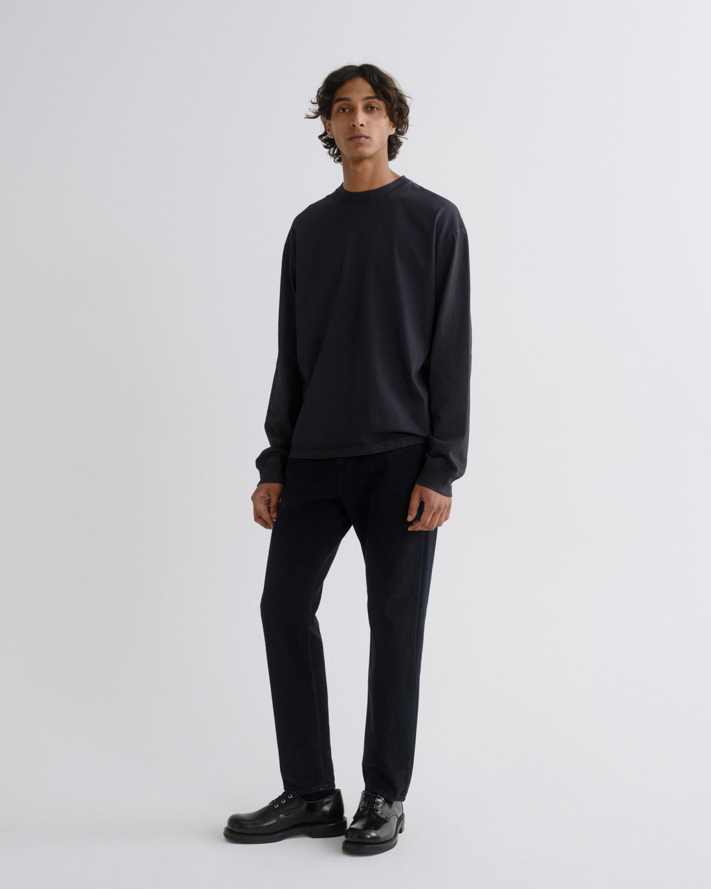 A man modeling a dark navy crewneck sweatshirt and matching dark navy trousers with black leather shoes, standing against a light gray studio background.
