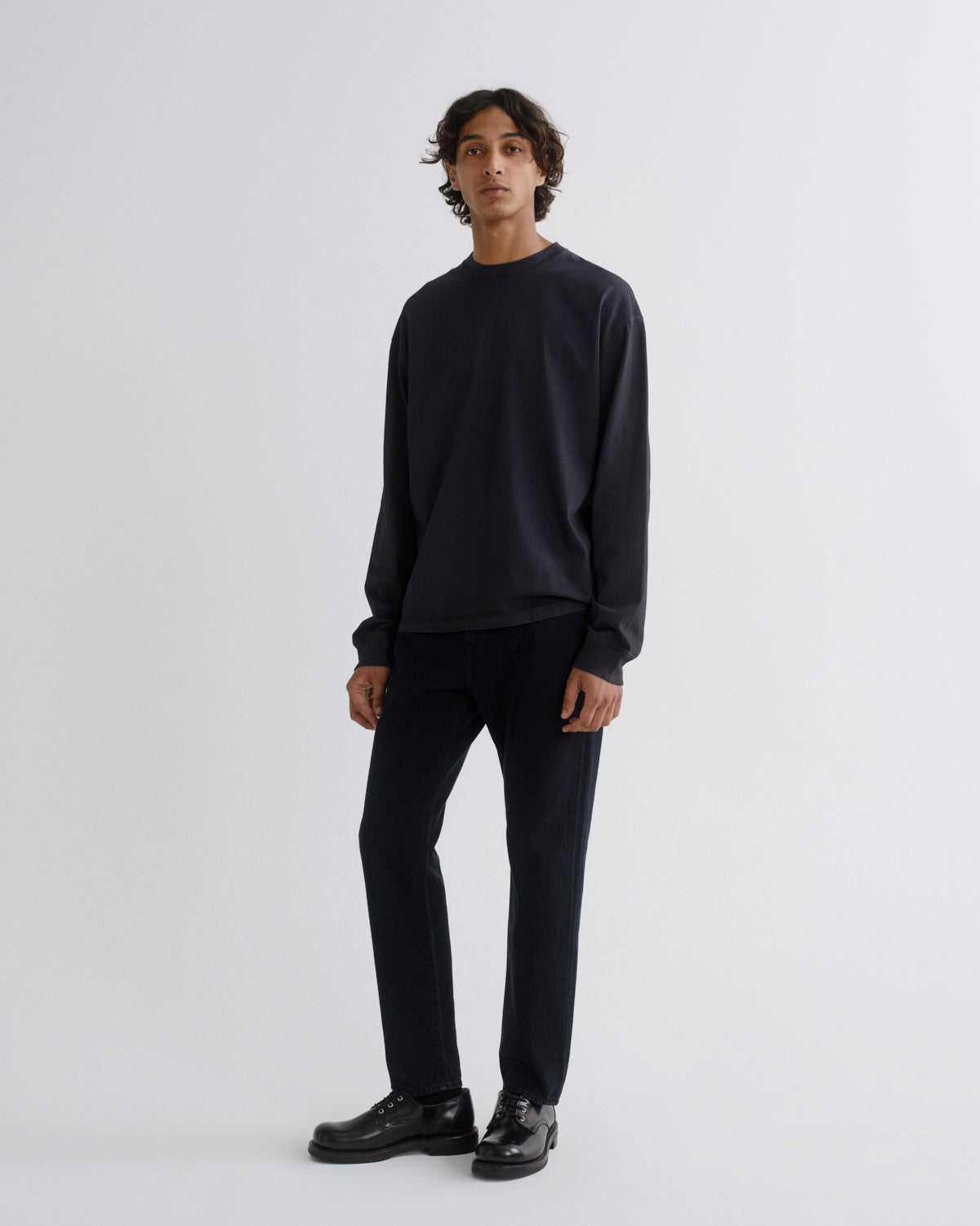 A man modeling a dark navy crewneck sweatshirt and matching dark navy trousers with black leather shoes, standing against a light gray studio background.