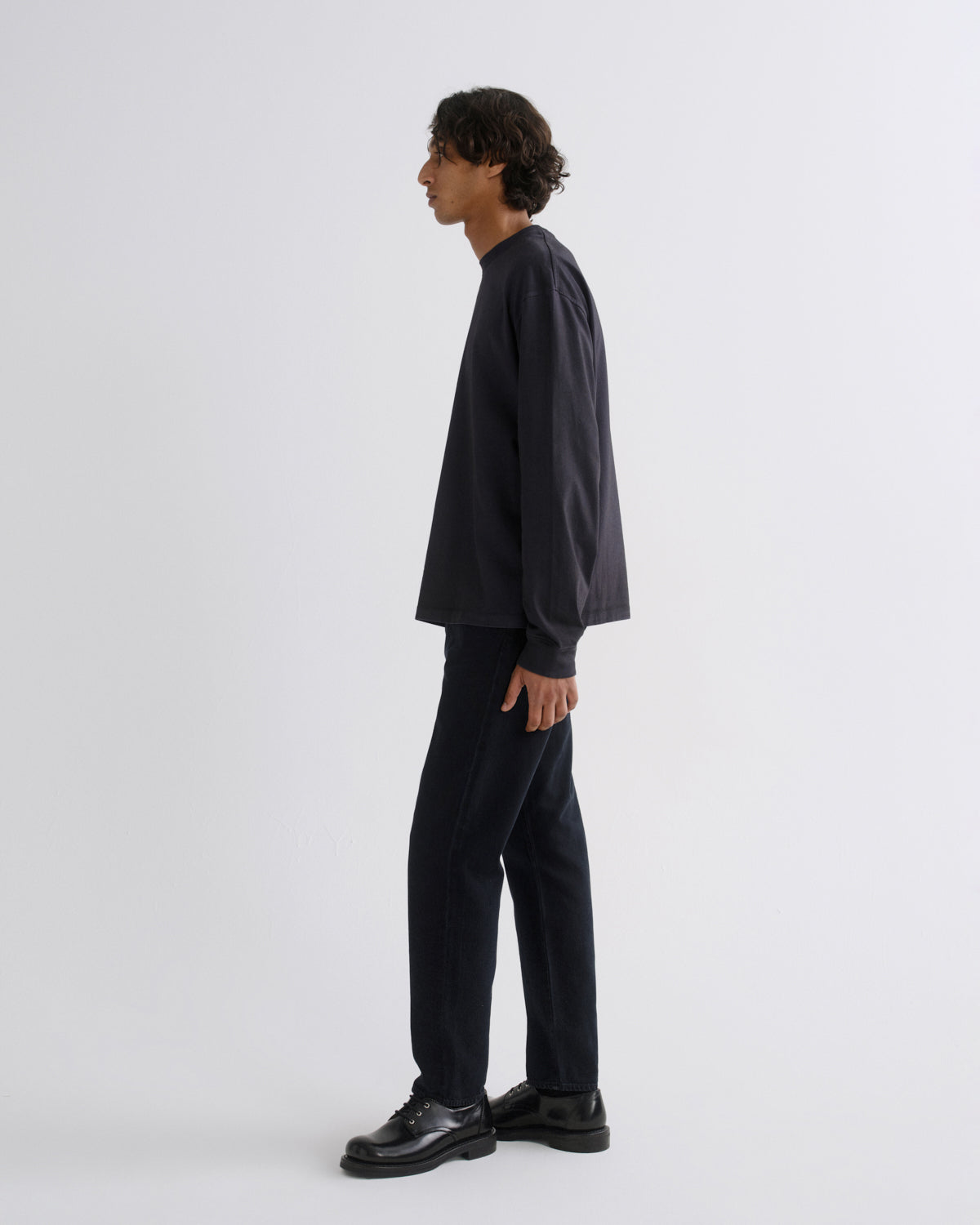A man in a dark navy long-sleeve shirt and matching dark trousers stands in profile against a light gray studio background.