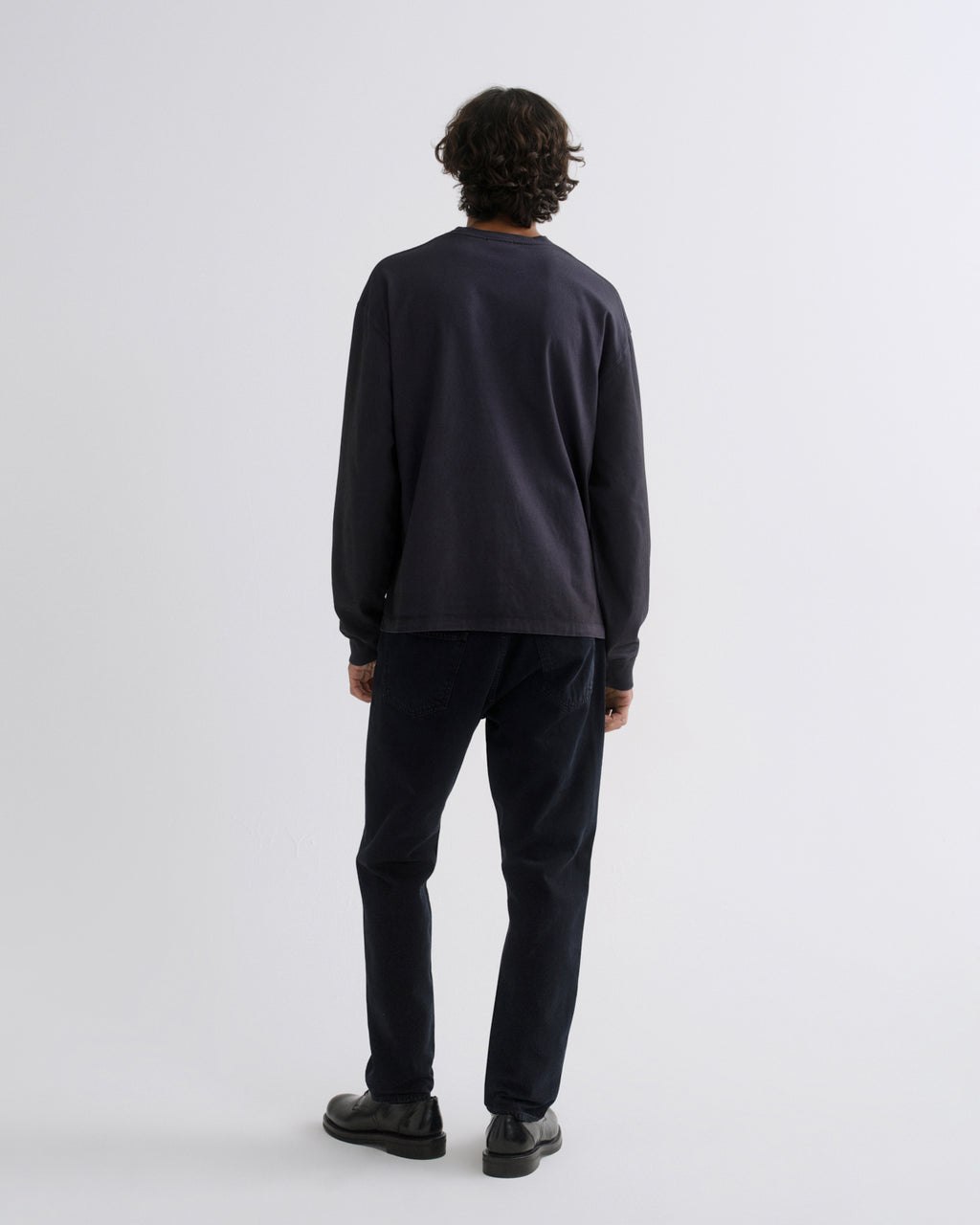 A man viewed from behind wearing a dark charcoal long-sleeve crewneck sweatshirt, dark navy pants, and black shoes, standing against a light gray studio background.