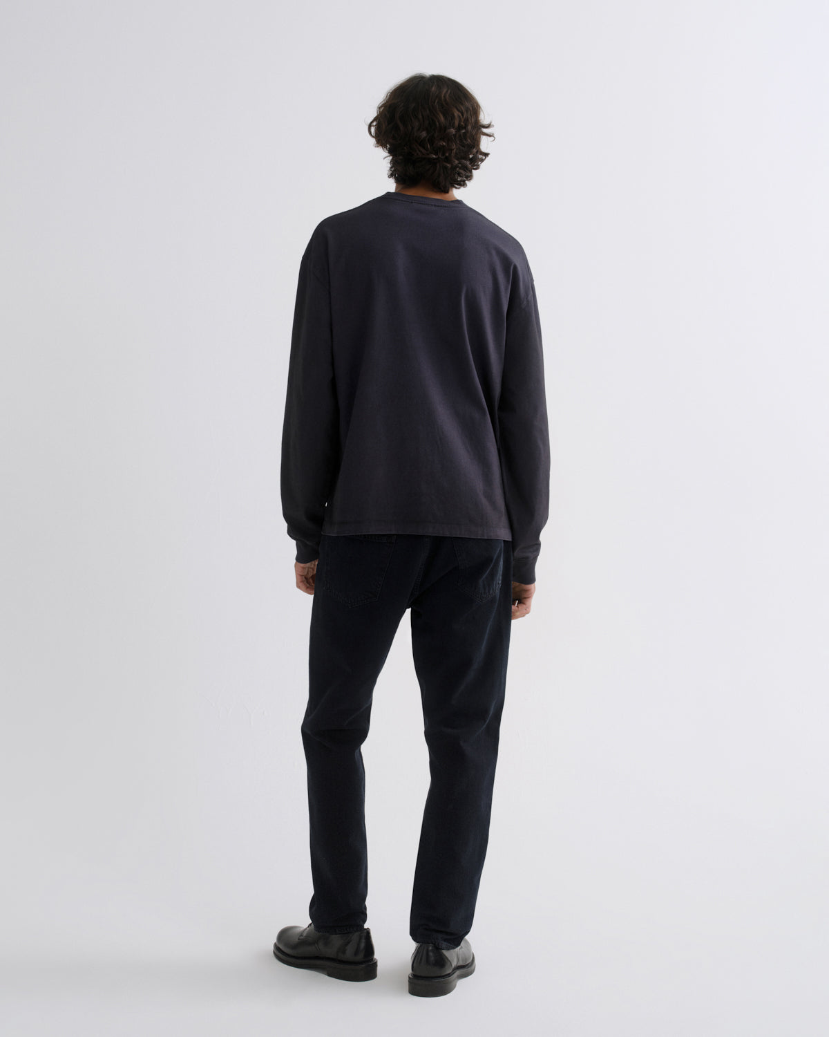 A man viewed from behind wearing a dark charcoal long-sleeve crewneck sweatshirt, dark navy pants, and black shoes, standing against a light gray studio background.