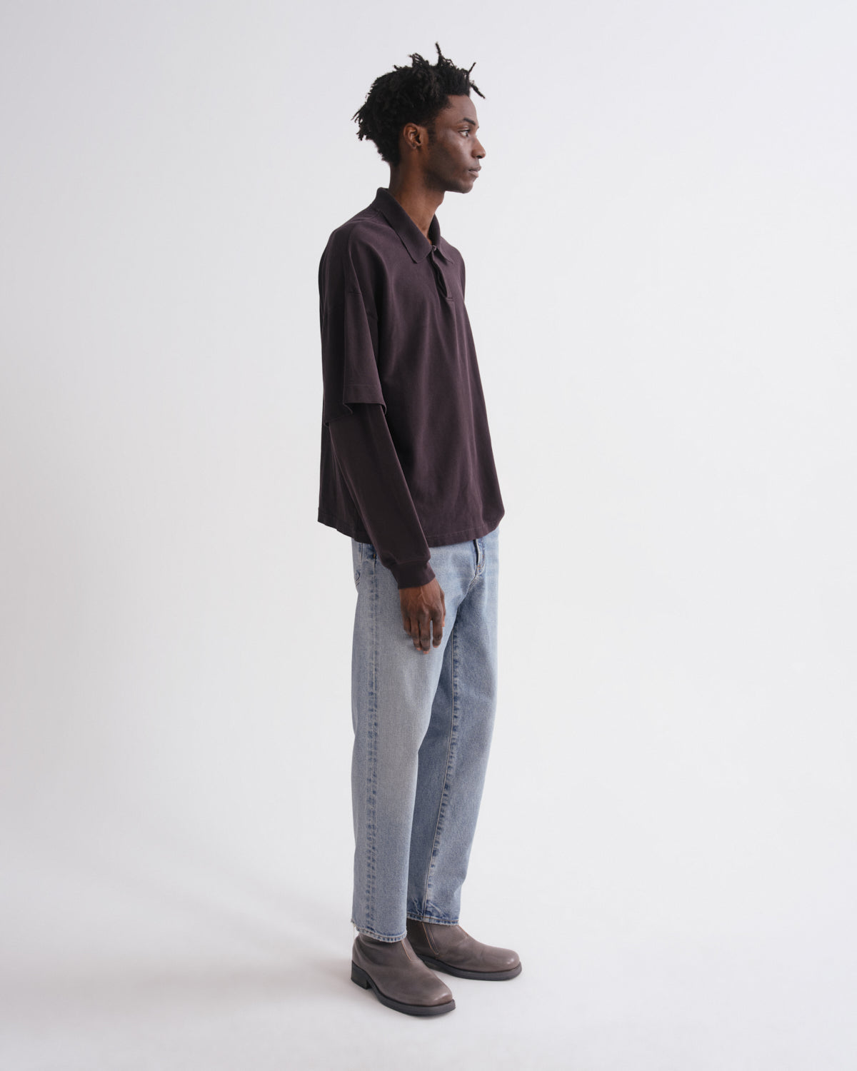 A man stands in profile wearing a dark plum button-up shirt, light-wash blue jeans, and brown suede boots against a plain white studio background.