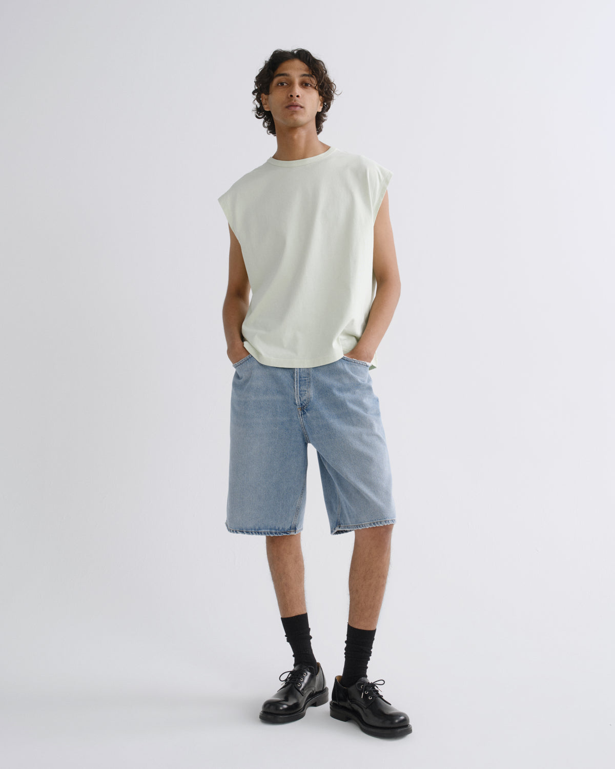 A model stands against a light gray studio background wearing a sleeveless pale green t-shirt, light-wash denim shorts, black socks, and black leather shoes.