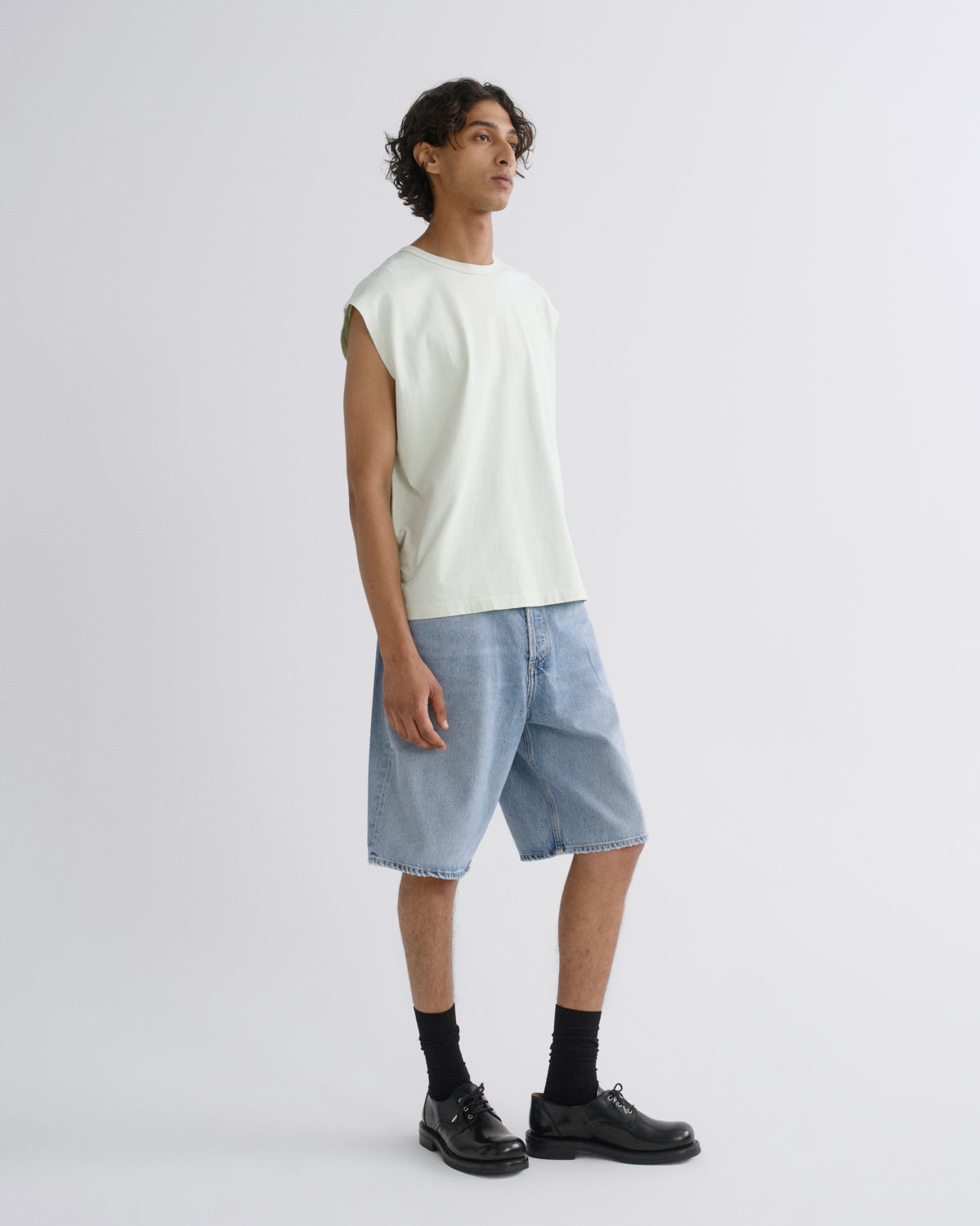 A man wearing a pale cream sleeveless t-shirt, light-wash denim shorts, black socks, and black leather shoes, standing against a light gray studio background.