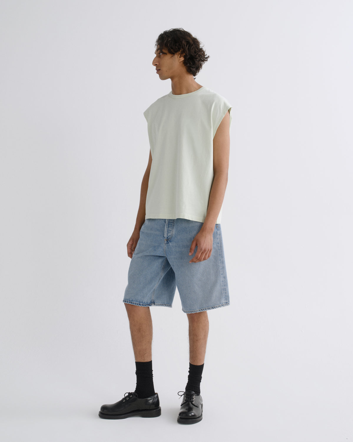 A model stands in profile wearing a light sage green sleeveless t-shirt, light-wash blue denim shorts, black crew socks, and black leather shoes against a plain light gray studio background.