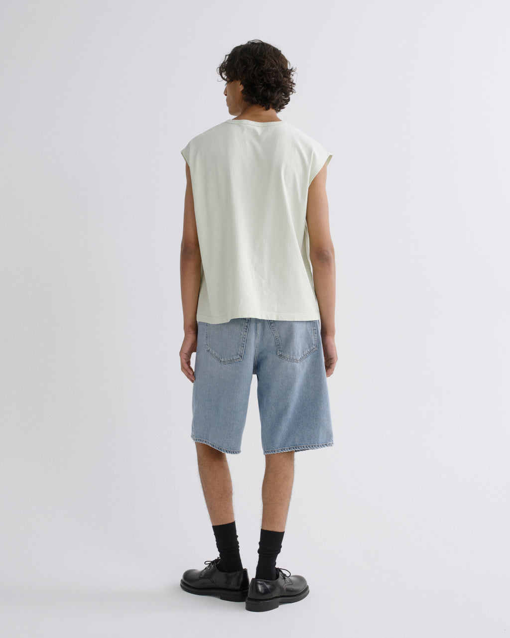 A model shown from the back wearing a light sage green sleeveless t-shirt, light blue denim shorts, black socks, and black leather shoes against a plain light gray background.