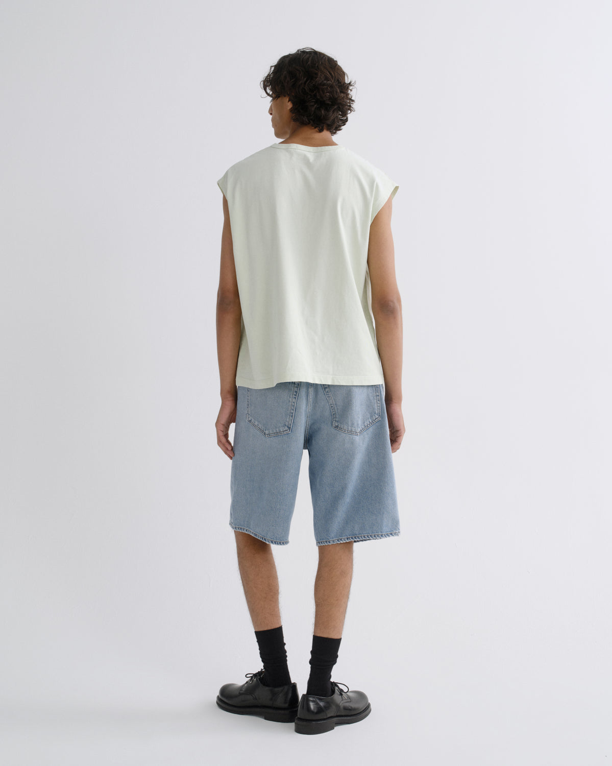 A model shown from the back wearing a light sage green sleeveless t-shirt, light blue denim shorts, black socks, and black leather shoes against a plain light gray background.