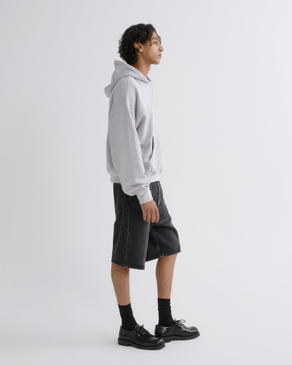A model shown in profile wearing a light gray pullover hoodie, dark gray denim shorts, black socks, and black leather shoes against a plain light gray studio background.