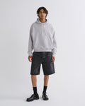 A model stands against a light gray studio background wearing a light gray pullover hoodie, dark gray denim shorts, black socks, and black leather shoes.
