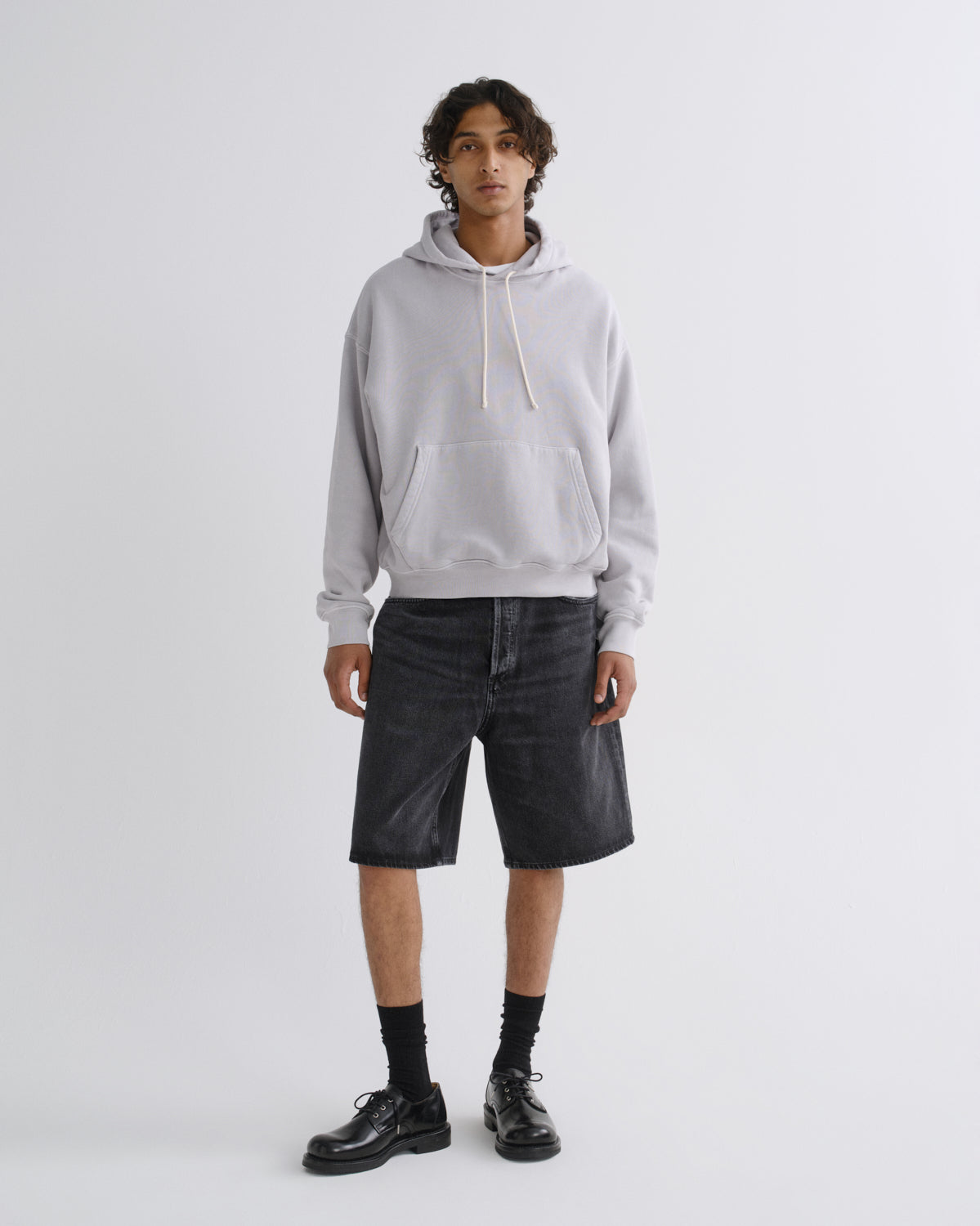 A model stands against a light gray studio background wearing a light gray pullover hoodie, dark gray denim shorts, black socks, and black leather shoes.