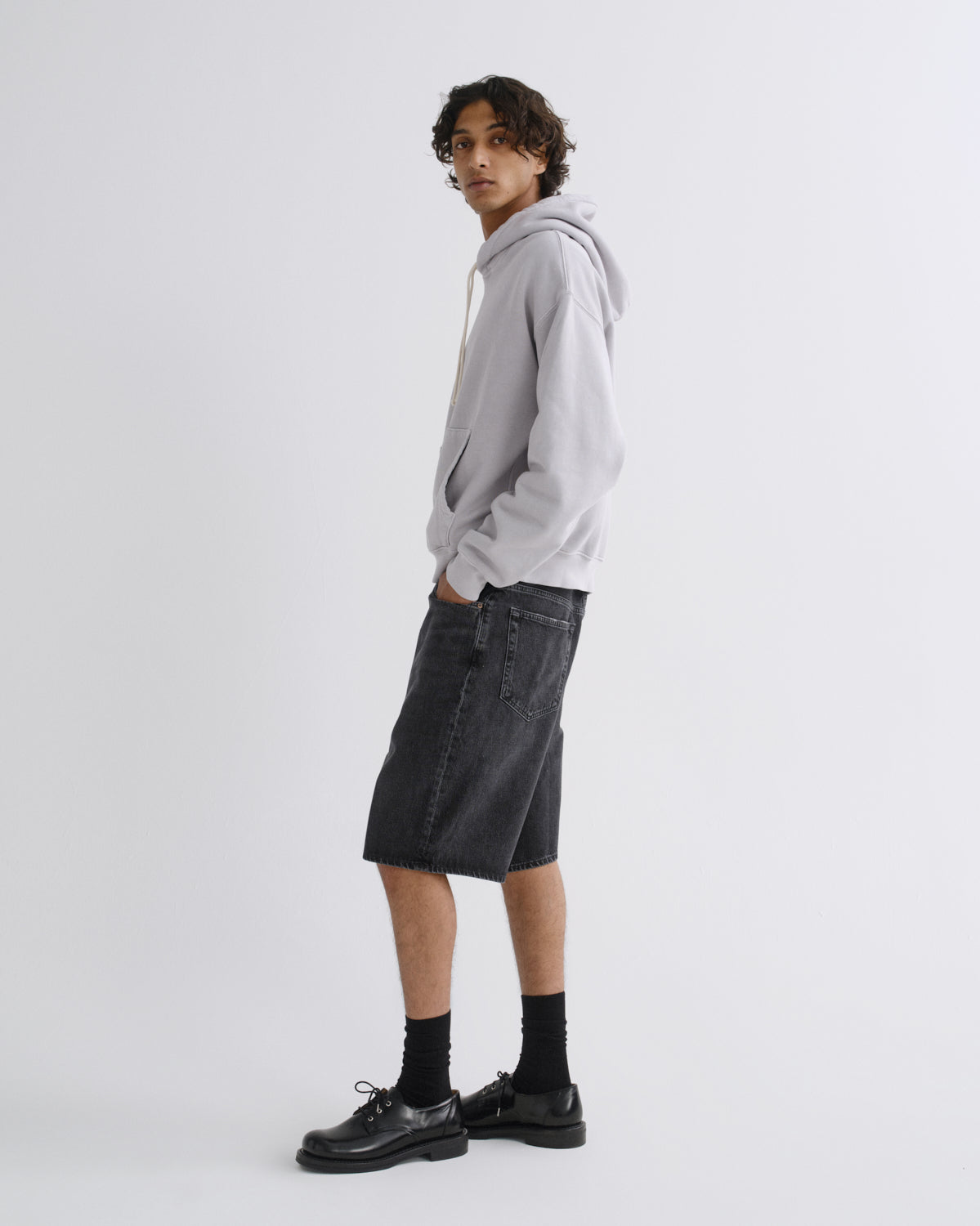 A model wearing a light gray oversized hoodie, dark gray denim shorts, black socks, and black leather shoes, standing in profile against a light gray studio background.