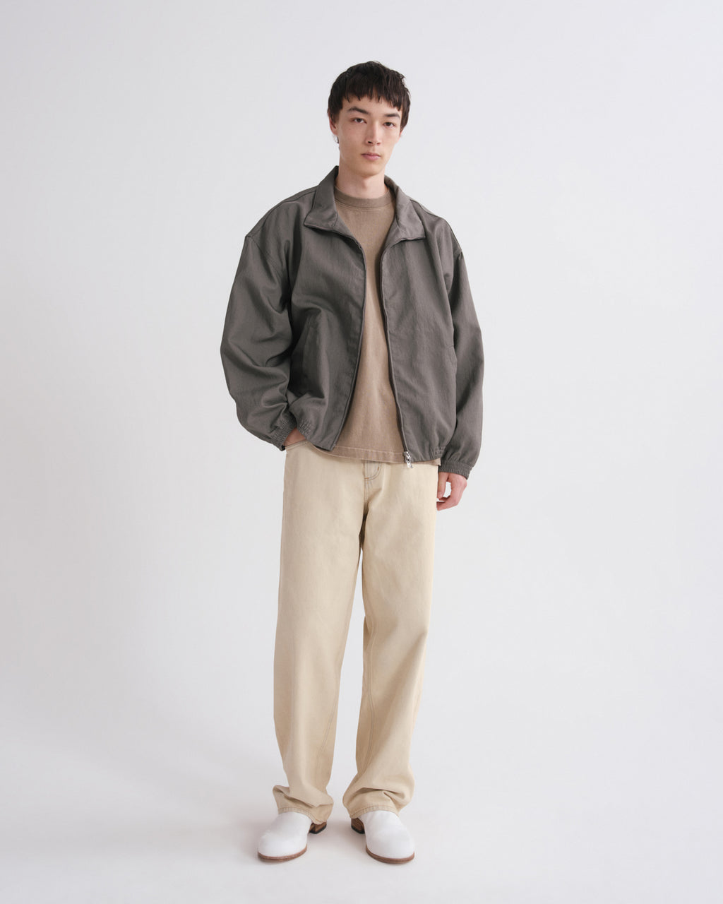 A model stands against a light gray studio background wearing a dark gray zip-up jacket over a beige turtleneck, paired with beige trousers and white shoes.