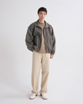 A model stands against a light gray studio background wearing a dark gray zip-up jacket over a beige turtleneck, paired with beige trousers and white shoes.
