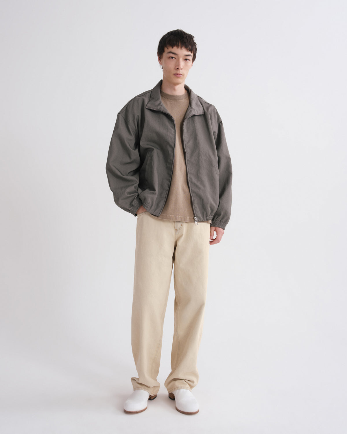 A model stands against a light gray studio background wearing a dark gray zip-up jacket over a beige turtleneck, paired with beige trousers and white shoes.