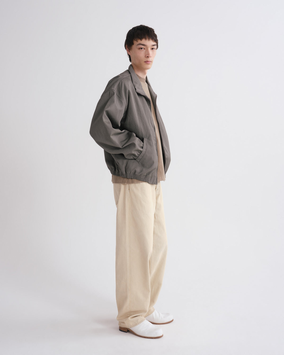 A model stands in profile wearing a dark gray bomber jacket over a beige knit top, paired with wide-leg beige trousers and white loafers, against a plain light gray studio background.