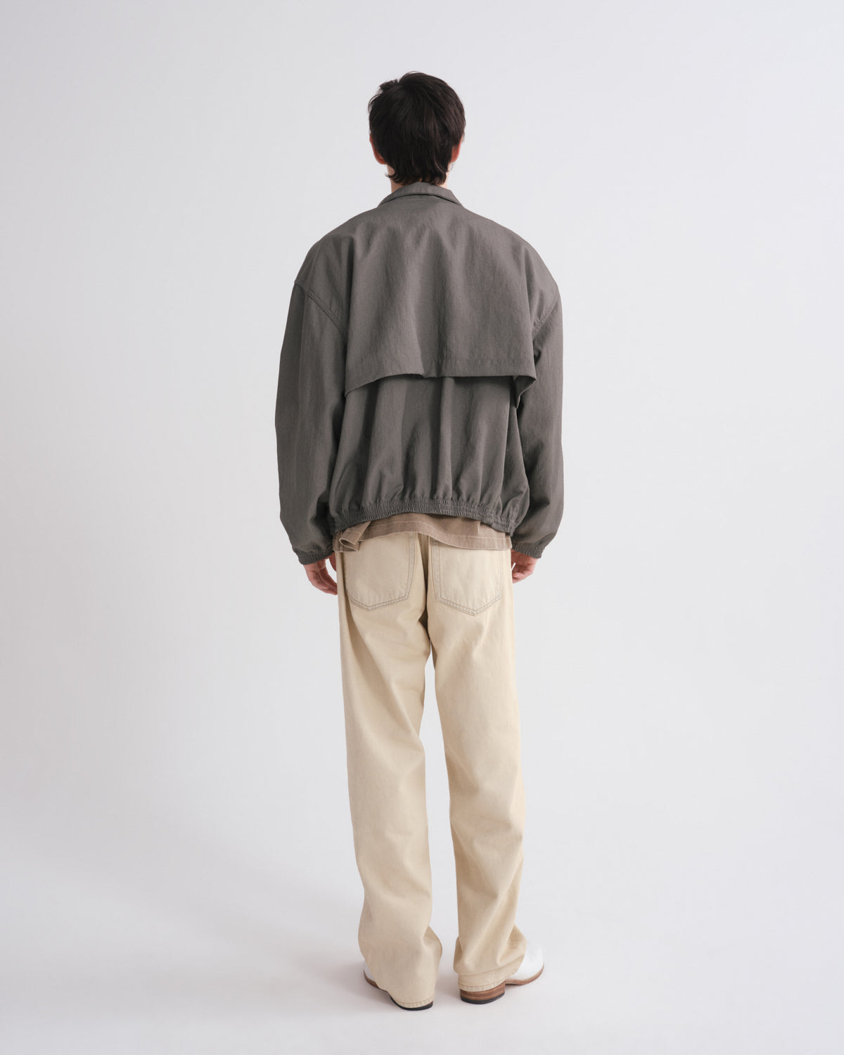 A person shown from the back wearing a dark gray oversized jacket and light beige trousers, standing against a plain light gray studio background.