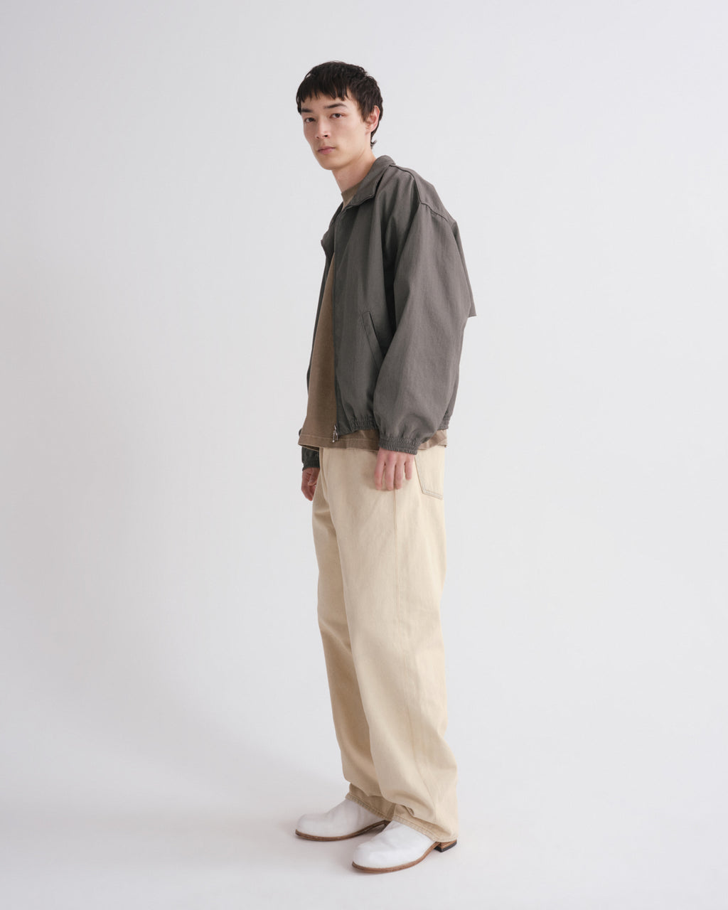 A man stands in profile wearing a dark gray oversized jacket over a tan shirt, cream wide-leg trousers, and white shoes against a plain light gray studio background.