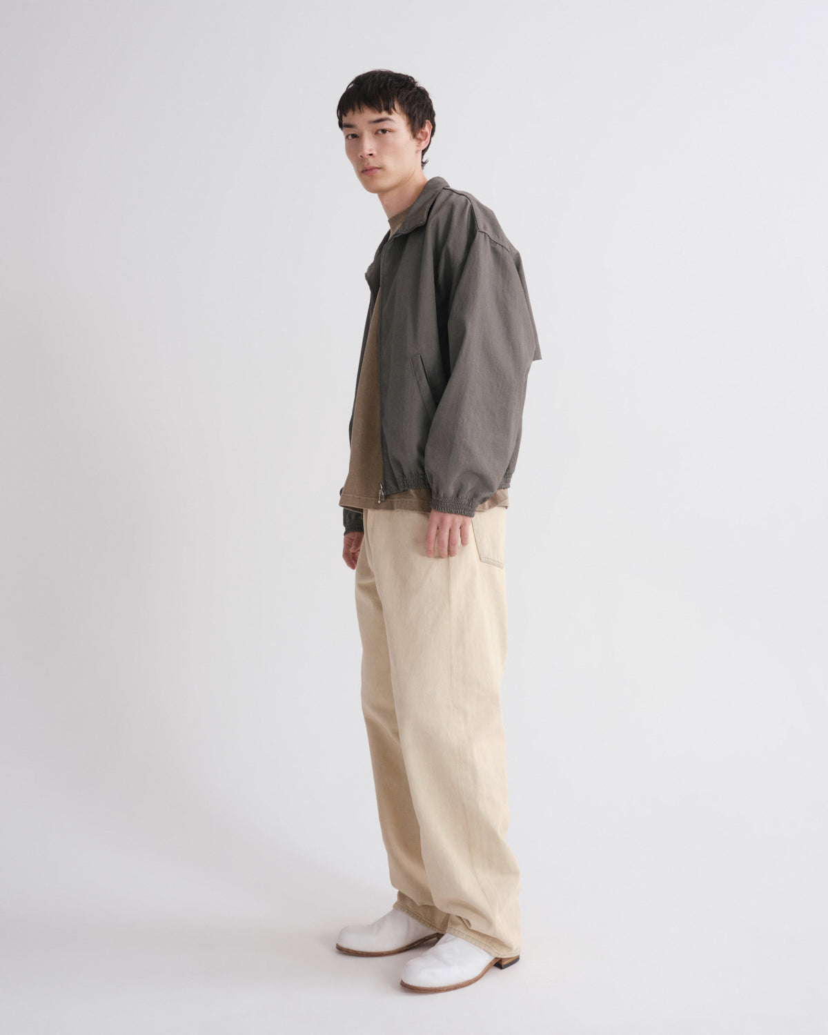 A man stands in profile wearing a dark gray oversized jacket over a tan shirt, cream wide-leg trousers, and white shoes against a plain light gray studio background.