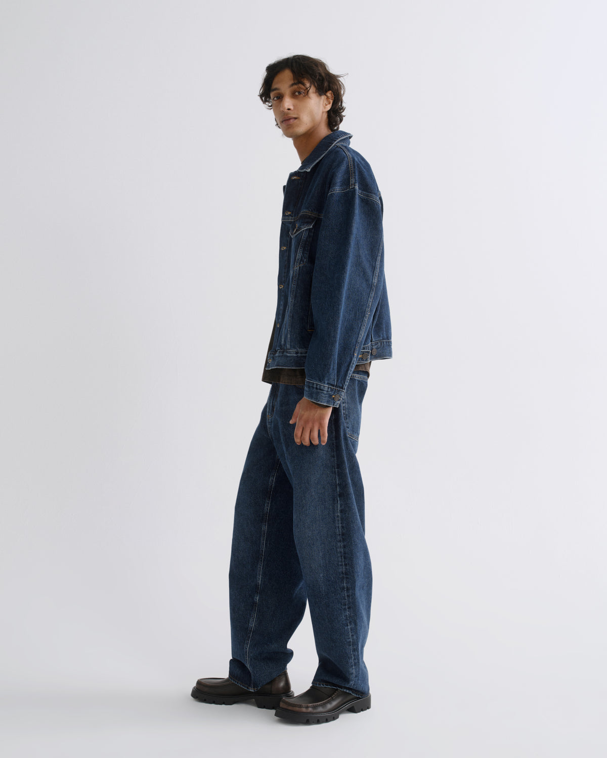 A model wearing a dark blue denim jacket and matching wide-leg denim jeans, paired with dark brown leather shoes, standing in profile against a light gray studio background.