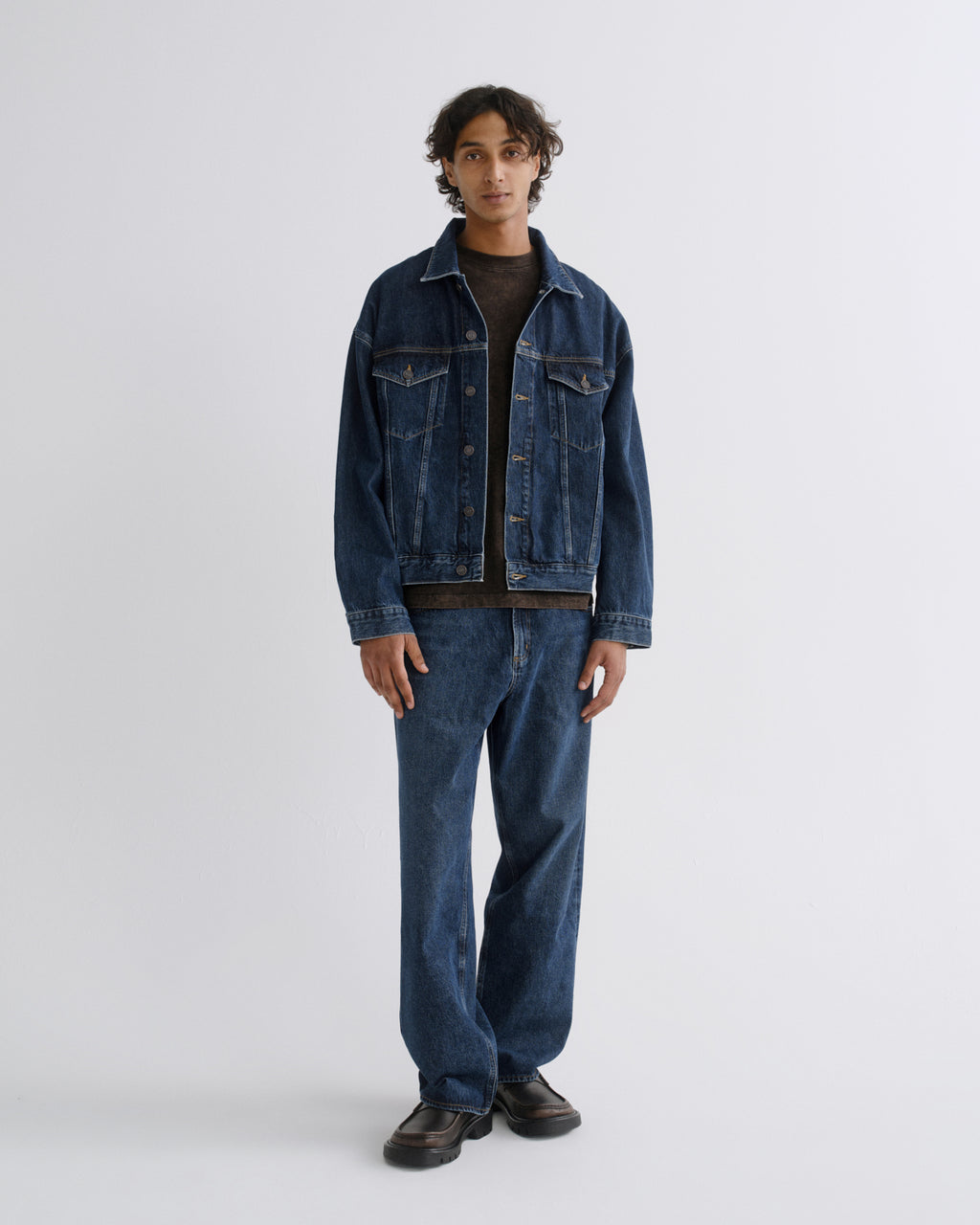 A model stands against a light gray studio background wearing a dark blue denim jacket and matching dark blue denim jeans over a black shirt and dark brown shoes.