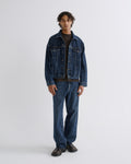 A model stands against a light gray studio background wearing a dark blue denim jacket and matching dark blue denim jeans over a black shirt and dark brown shoes.