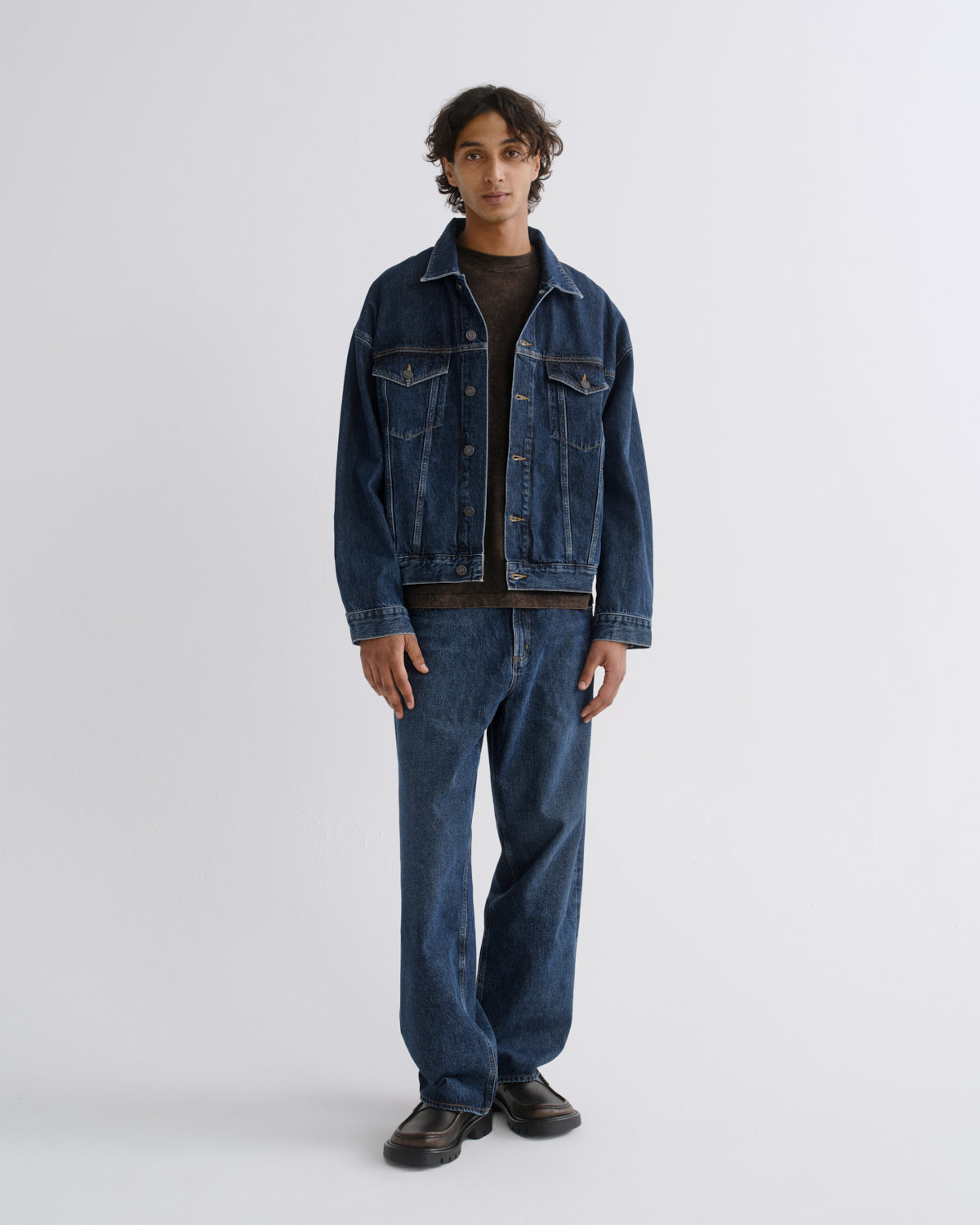 A model stands against a light gray studio background wearing a dark blue denim jacket and matching dark blue denim jeans over a black shirt and dark brown shoes.