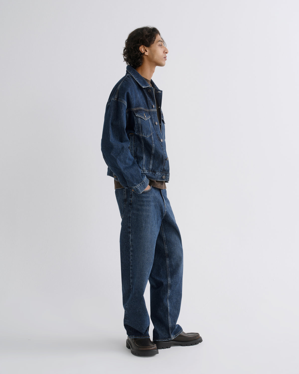 A model in side profile wearing a dark blue denim jacket and matching dark blue wide-leg jeans, standing against a light gray studio background.