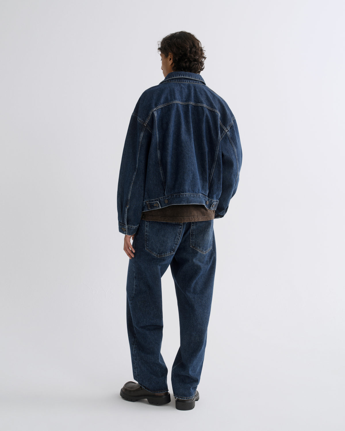 A man viewed from behind wearing a dark blue denim jacket and matching dark blue jeans, standing against a plain light gray studio background.