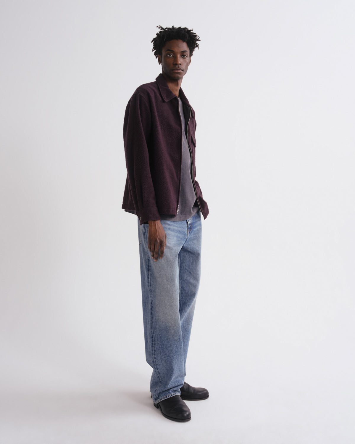 Man wearing a burgundy shirt, gray t-shirt, and blue jeans on a white background