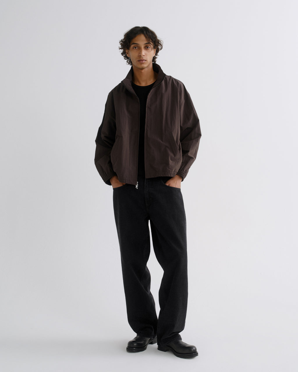 A model stands against a light gray studio background wearing a dark brown lightweight jacket over a black shirt and black wide-leg trousers.
