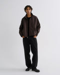 A model stands against a light gray studio background wearing a dark brown lightweight jacket over a black shirt and black wide-leg trousers.