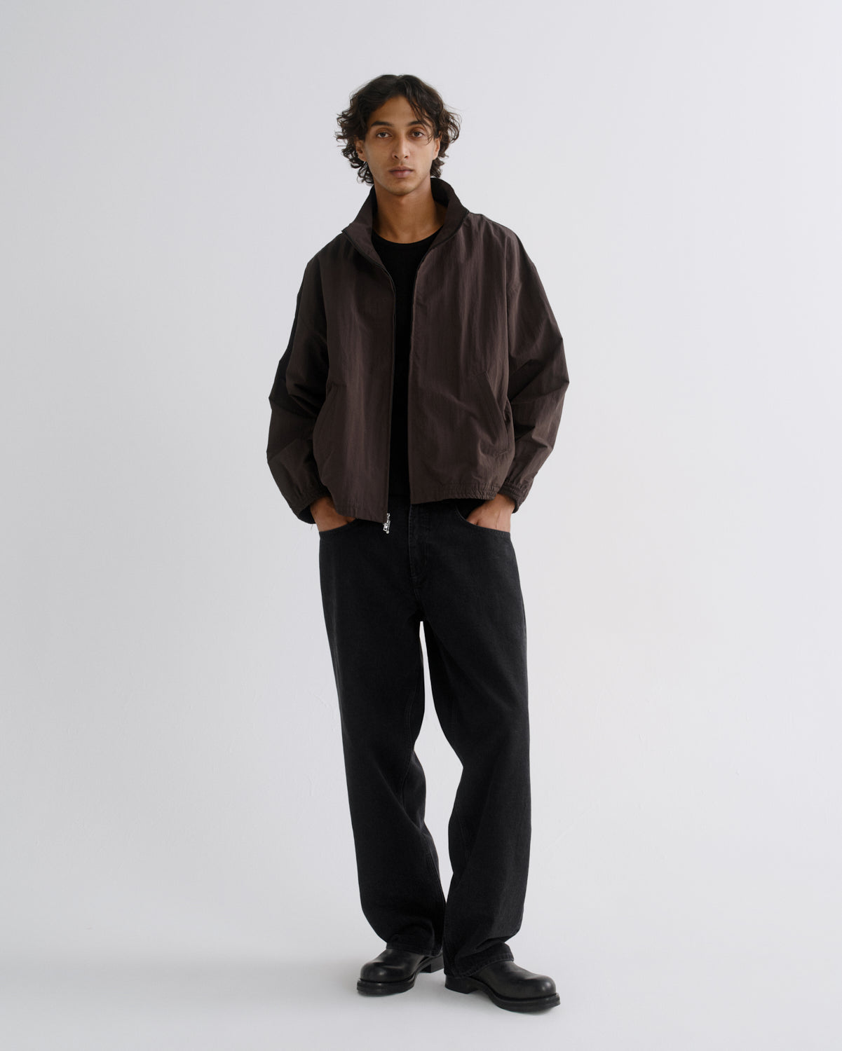 A model stands against a light gray studio background wearing a dark brown lightweight jacket over a black shirt and black wide-leg trousers.