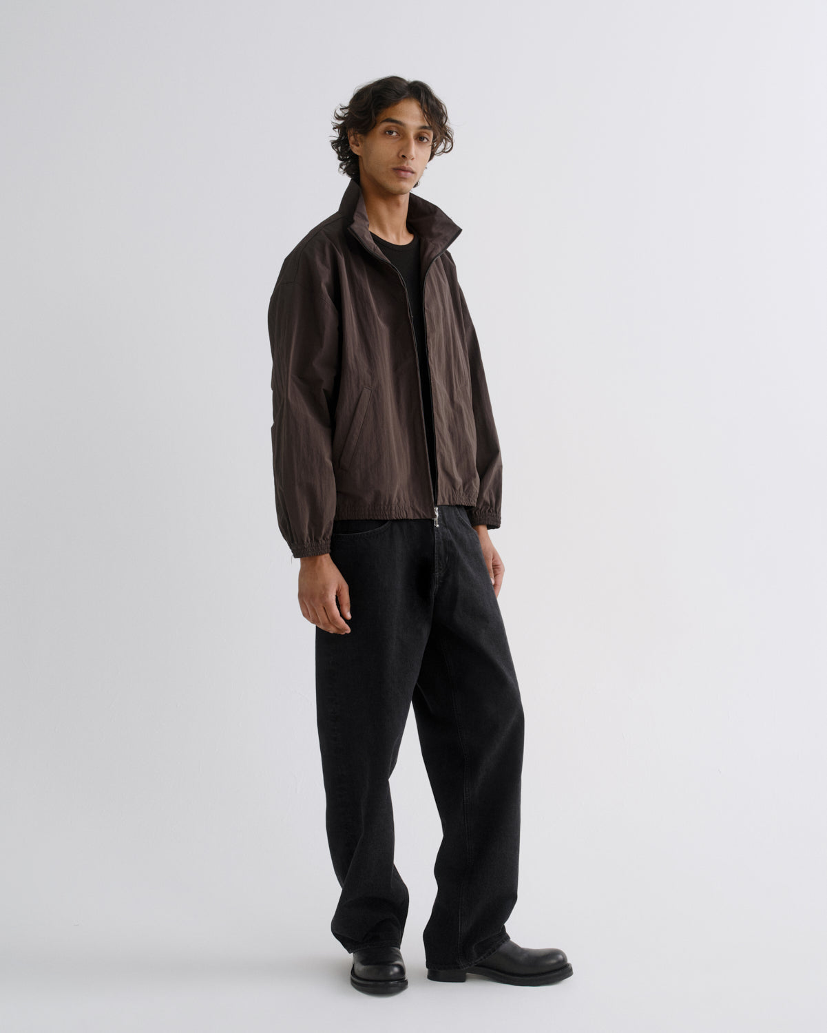 A model stands in a studio wearing a dark brown lightweight zip-up jacket over a black shirt and black wide-leg trousers.