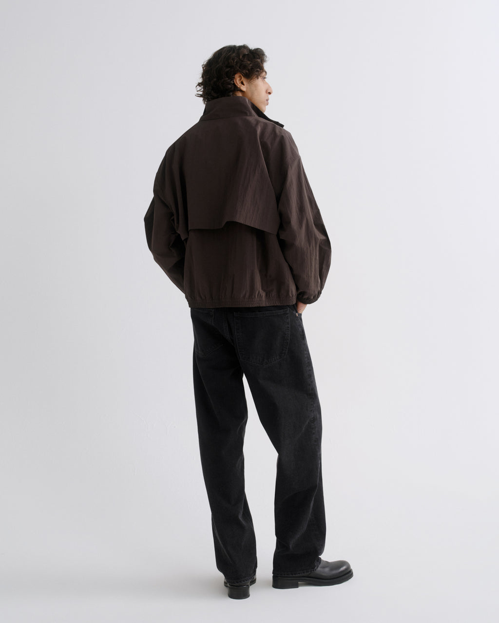 A man is shown from the back wearing a dark brown utility jacket and black pants against a light gray studio background.