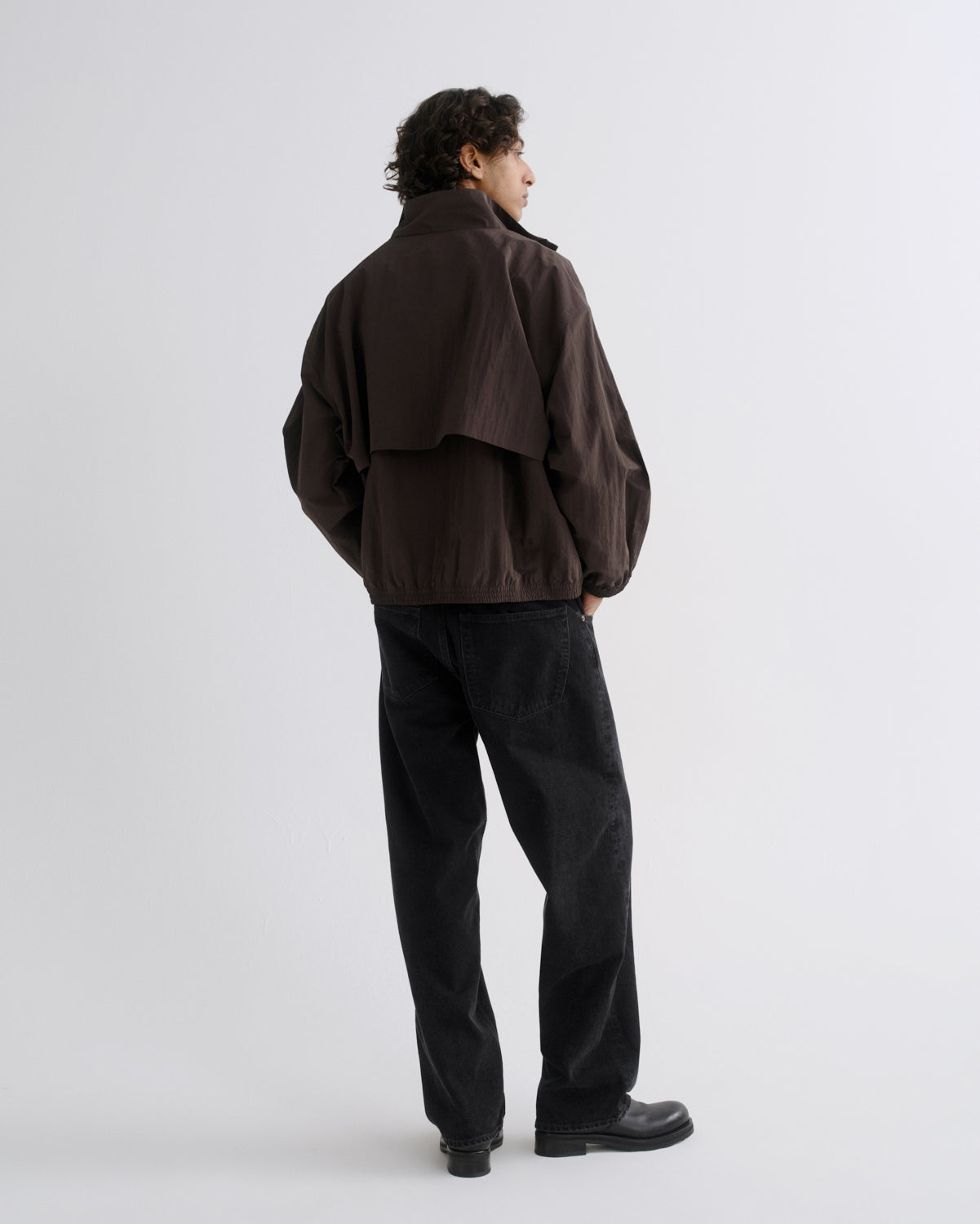 A man is shown from the back wearing a dark brown utility jacket and black pants against a light gray studio background.