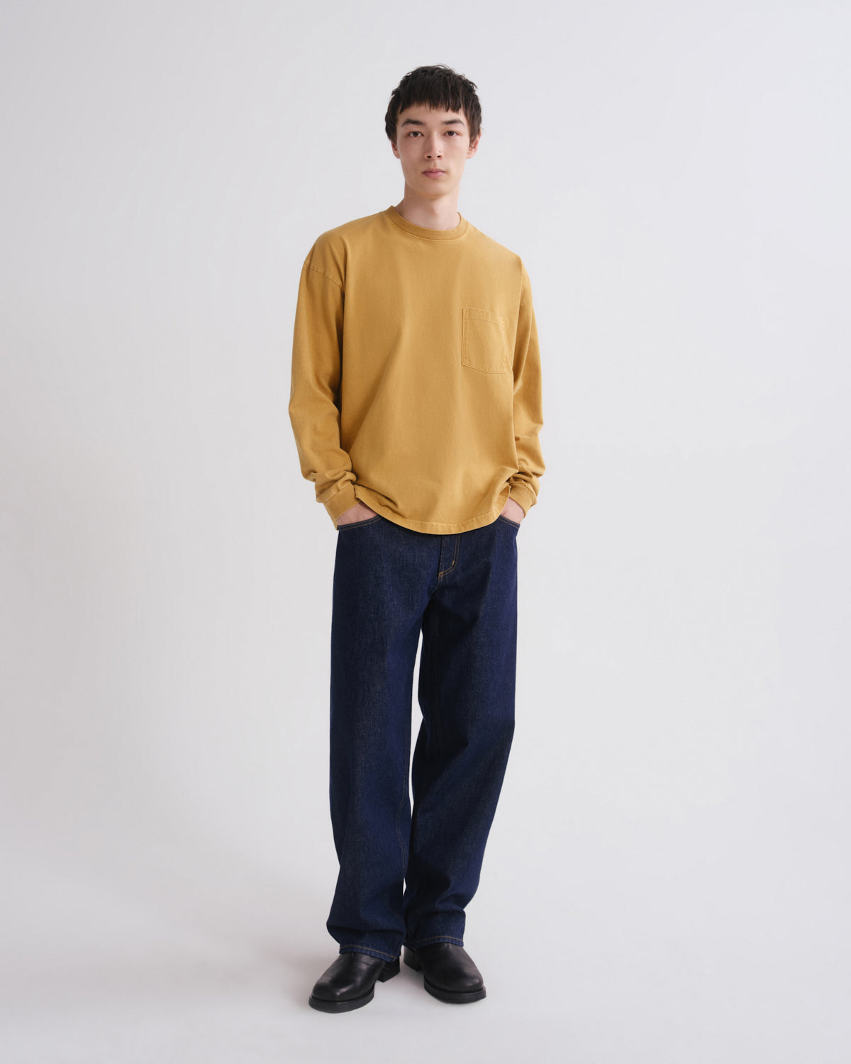 A model stands against a light gray studio background wearing a mustard yellow long-sleeve crewneck shirt with a chest pocket, dark blue jeans, and black shoes.