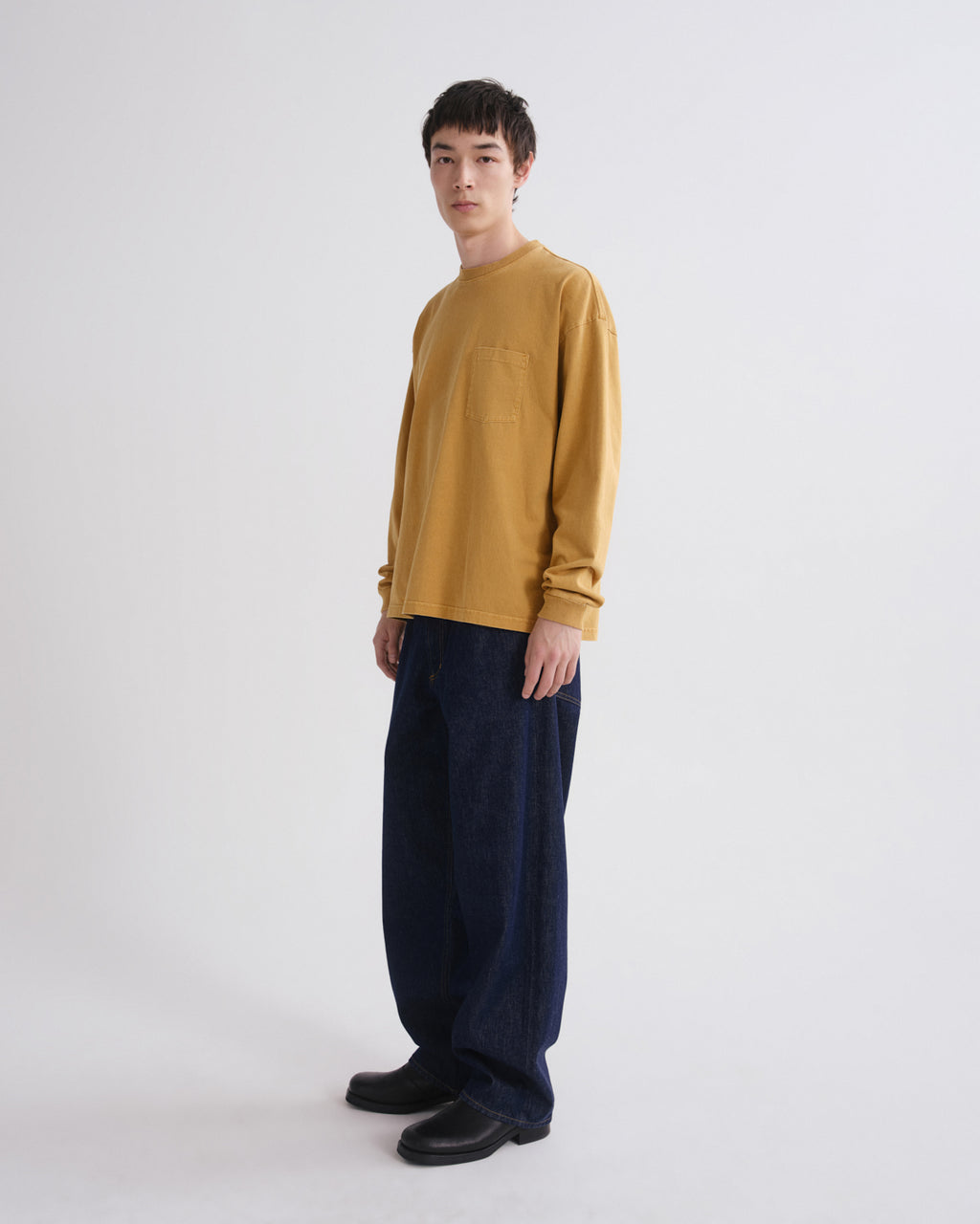 A man wearing a mustard yellow long-sleeve crewneck sweater and dark navy wide-leg trousers, standing against a plain light gray studio background.