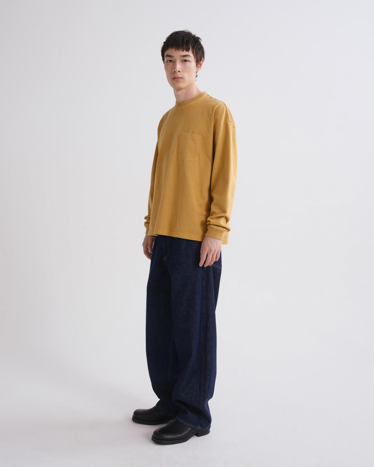 A man wearing a mustard yellow long-sleeve crewneck sweater and dark navy wide-leg trousers, standing against a plain light gray studio background.