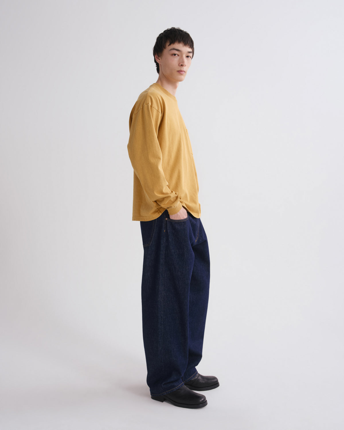 A model standing in profile wearing a mustard yellow long-sleeve shirt, dark navy wide-leg trousers, and black shoes against a plain light gray studio background.