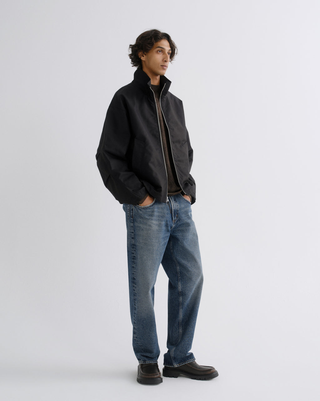 A man wearing a black zip-up jacket, blue jeans, and dark brown shoes, standing against a light gray studio background.